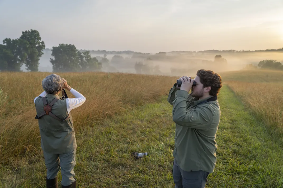 Photo of two people standing in a pasture looking through binoculars.