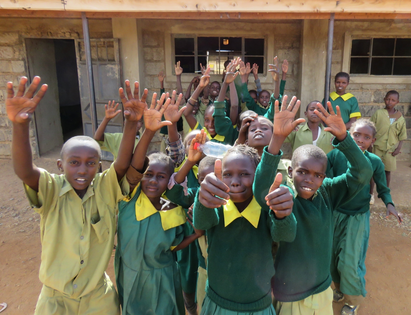 Primary school students in Laikipia, Kenya, pose for a photo in front of a building. Some of the students are raising their hands and giving a thumbs up