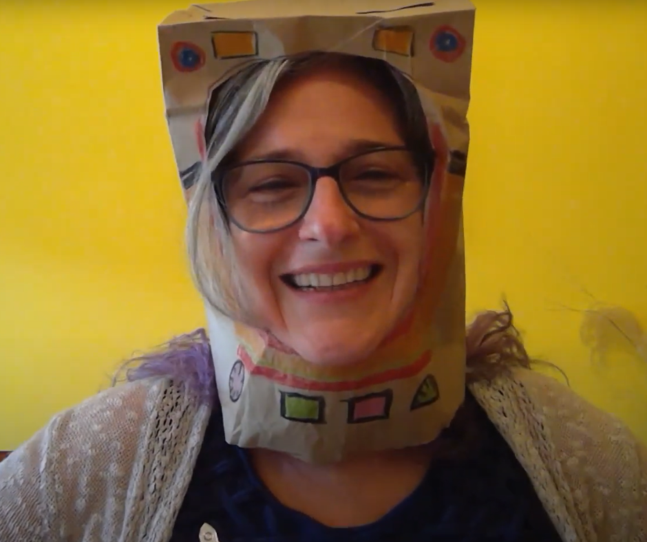 A woman smiles while wearing a paper bag astronaut helmet in front of a bright yellow background.