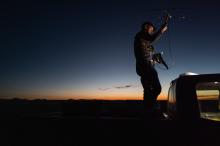 A nighttime photo of a man standing on the back of a pickup truck. He is using a satellite antenna to track wild ferrets. 