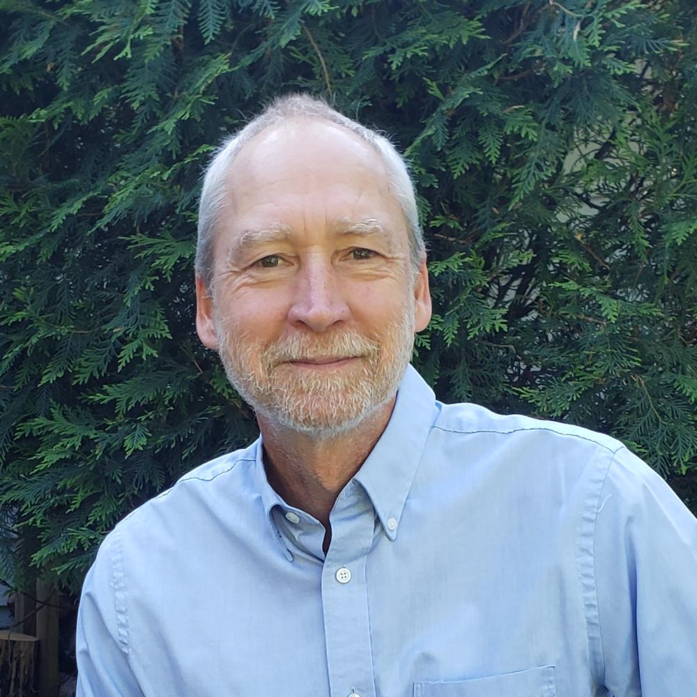 A man with white hair and a blue oxford shirt smiles in front of a leafy green background.