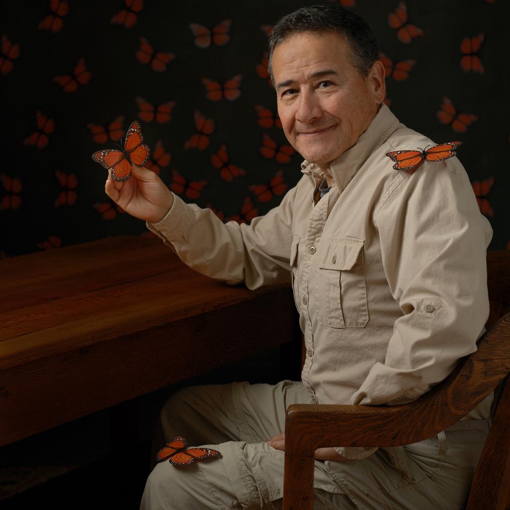 Portrait of man in a tan worksuit smiling at the camera as an orange butterfly perches on his finger.