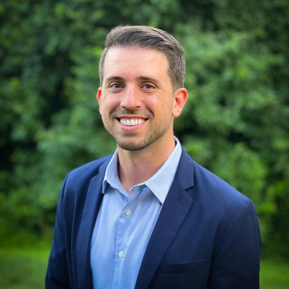 A photo of a man wearing a blue suit and lighter blue button down shirt standing in front of green foliage