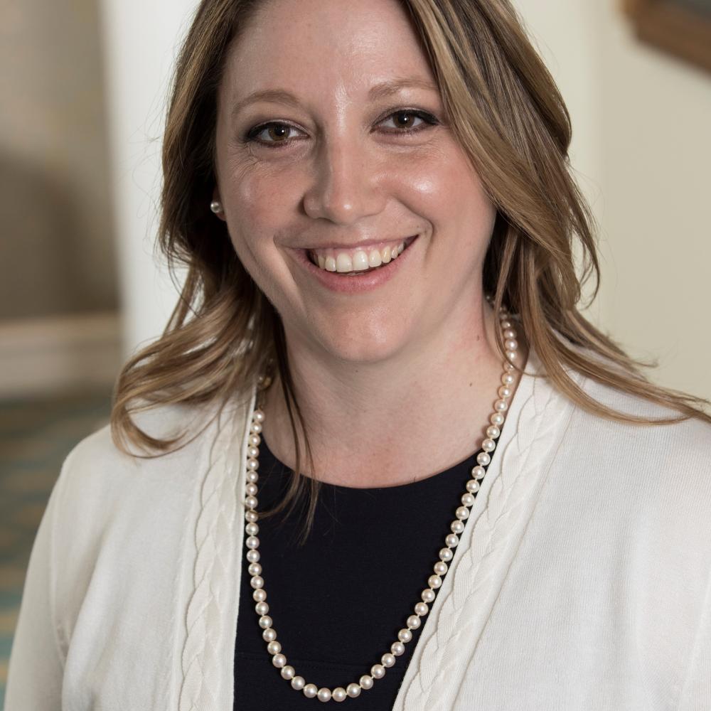 A photo of a woman with shoulder-length brown hair wearing a strand of long pearls over a black shirt and white sweather