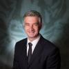 A gray-haired scientist wearing a black suit and tie.