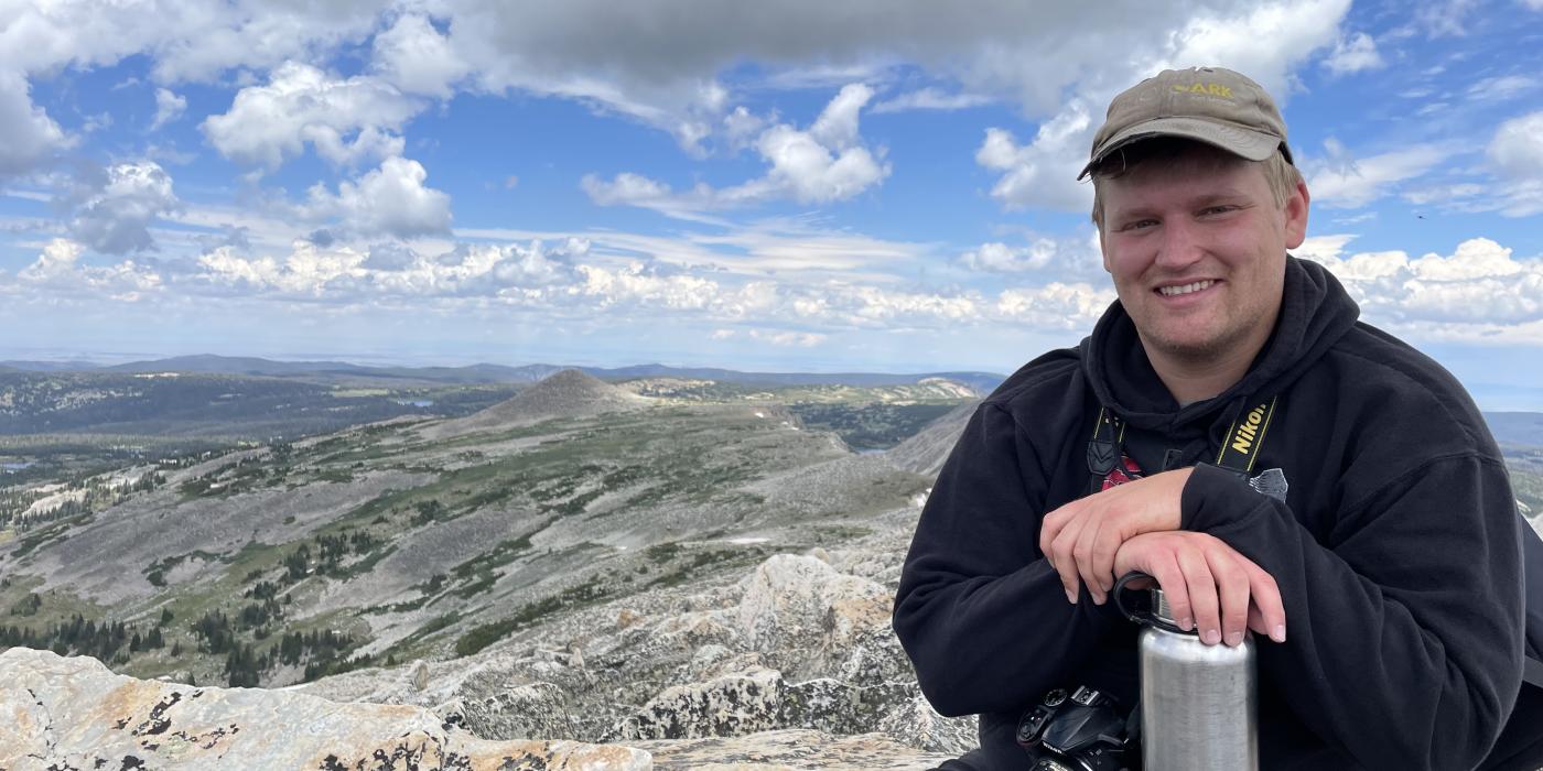 Photo of a young Caucasian man sitting on a mountain top, smiling at the camera.