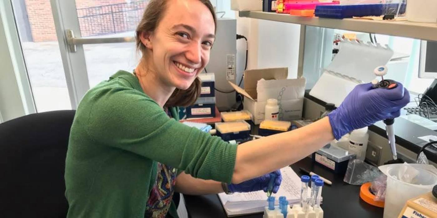 Photo of Alyssa Kaganer in a lab setting using a pipette. She is smiling toward the viewer.