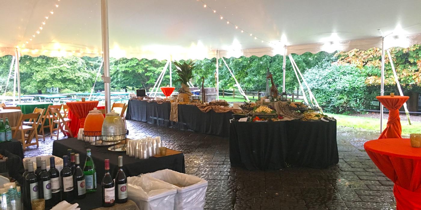 A bar and decorated tables set up under a large event tent lit up with lights in the Great Meadow