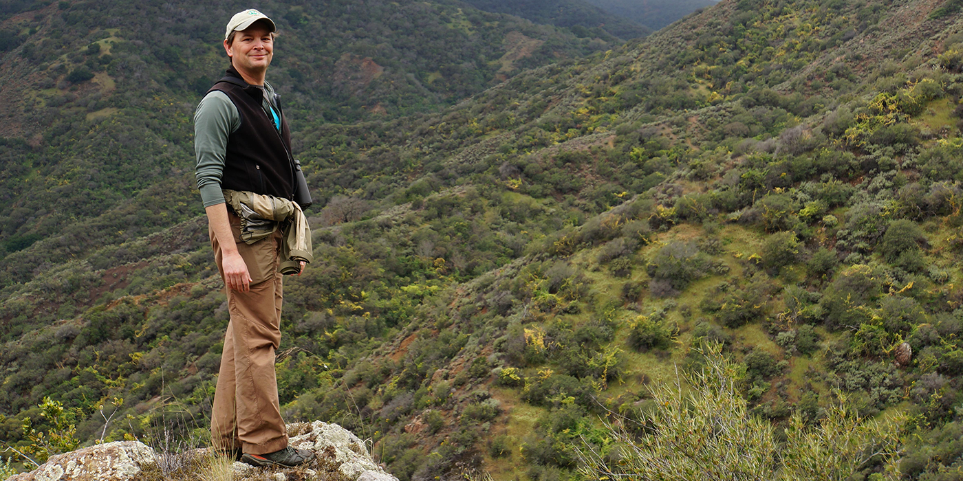 A man wearing a vest and khaki pants stands on a rock overlooking a green valley