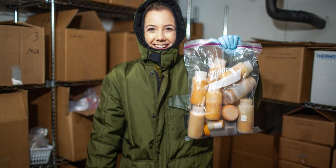 A nutrition lab research assistant at the Zoo wearing a full-body snowsuit stands in the freezer where animal milk samples are stored. She holds a bag full of milk samples, and is surrounded by shelves stacked with cardboard boxes full of more samples.